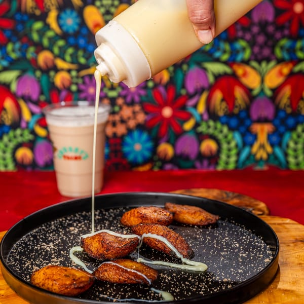 Hand squeezing a creamy sauce over fried plantains on a black plate with a drink in the background.