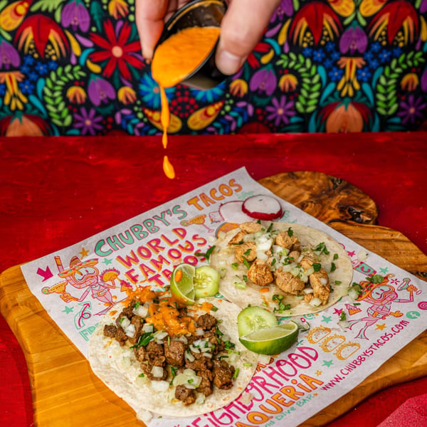 A hand pours orange sauce onto a Chubby's Tacos beef taco next to a chicken taco.
