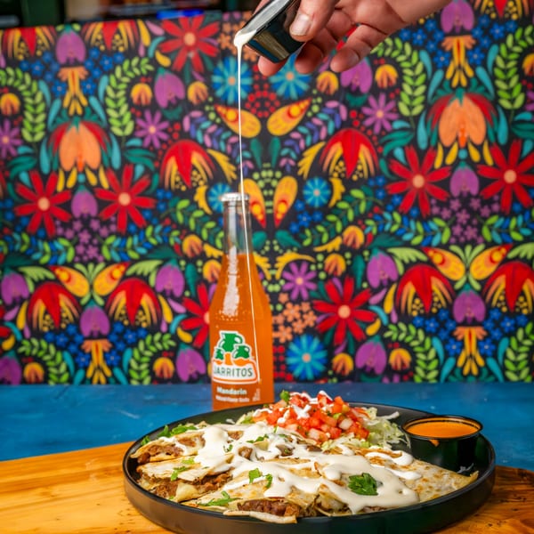 A hand drizzles white sauce over cheesy quesadillas with pico de gallo and a Jarritos soda.