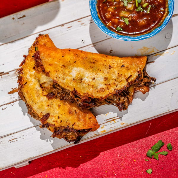 Two crispy birria tacos with shredded beef and cheese, served with a side of consommé.